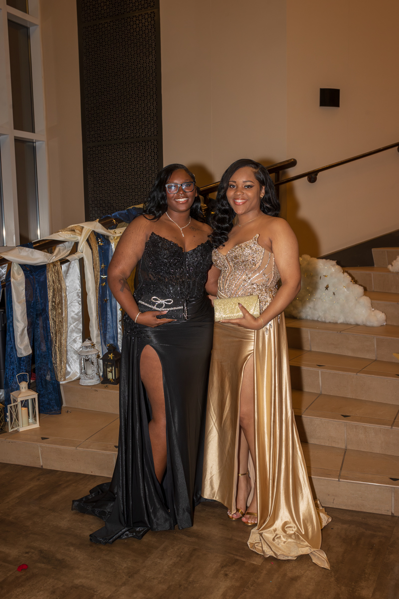 Two women in formal gowns posing together on a stair step at an event, smiling for the camera. One wears a black gown with a thigh-high slit; the other a gold beaded dress with a matching clutch.