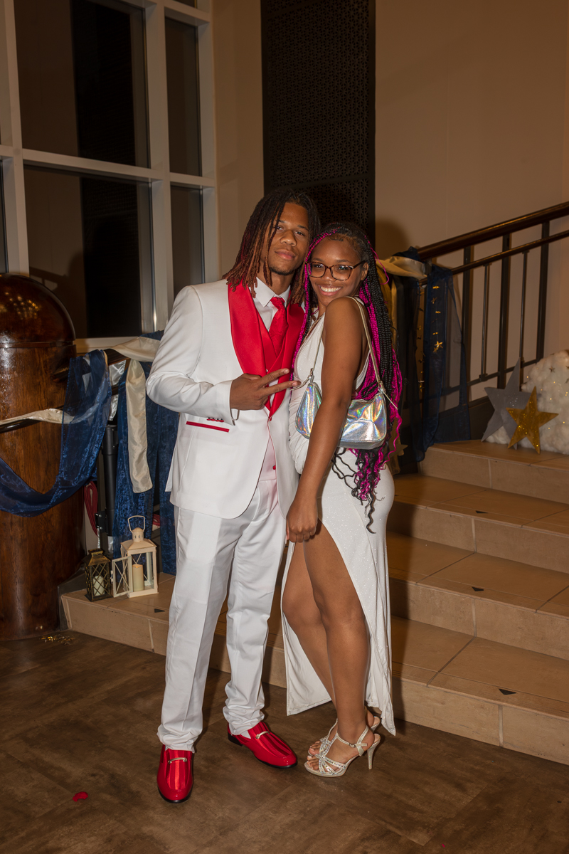 Couple wearing white formal outfits posing together indoors at a decorated staircase event, smiling at the camera.