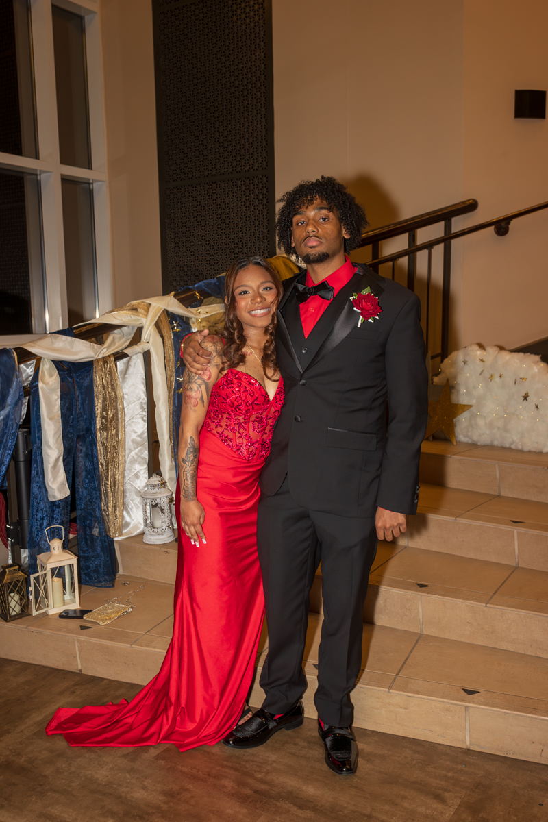 Couple dressed in formal wear posing on staircase; woman in red gown and man in black suit with red shirt and boutonniere.