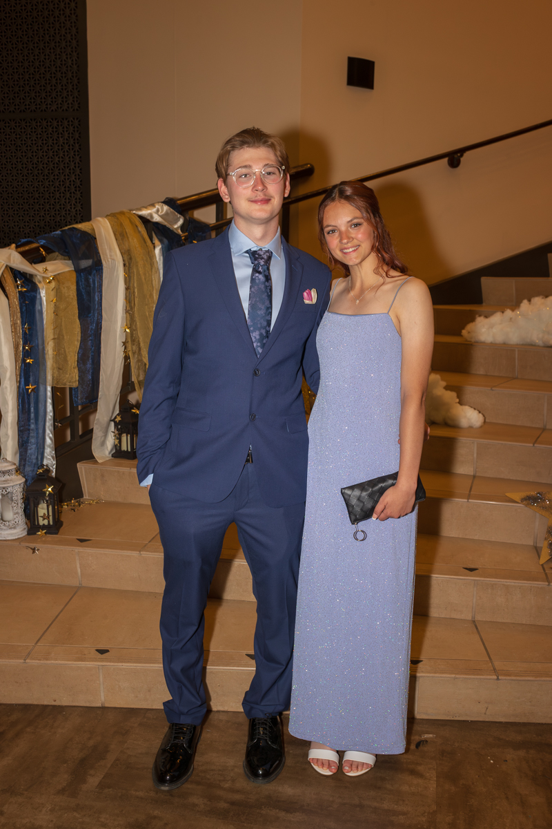 Young couple in formal attire posing on a decorated staircase; man in a blue suit, woman in a light blue glittery dress with a black clutch.