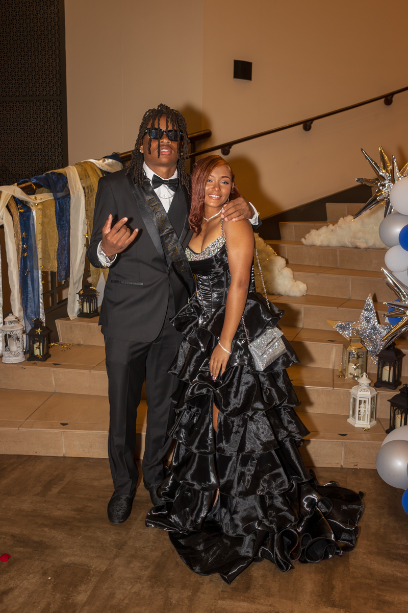 A couple in formal wear posing on steps: man in a black tuxedo with sunglasses and a bow tie, woman in a voluminous black gown smiling beside him.
