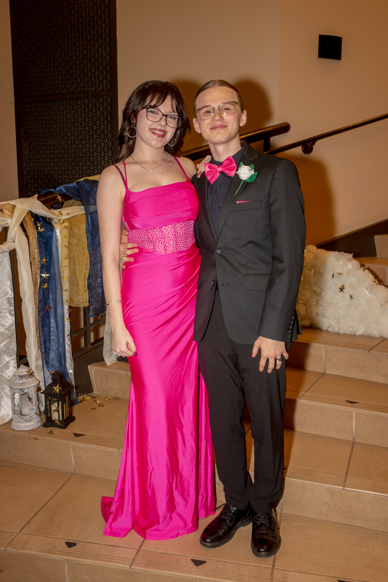 Two teens in formal wear posing on a staircase: girl in a bright pink gown and the boy in a black suit with a pink bow tie, smiling together at a formal event.