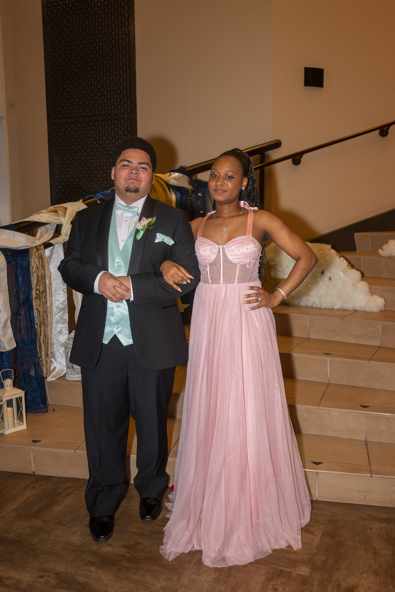 Couple in formal attire posing on stairs: man in black tux with mint vest and boutonniere, woman in pink strapless-style gown with jeweled bodice.