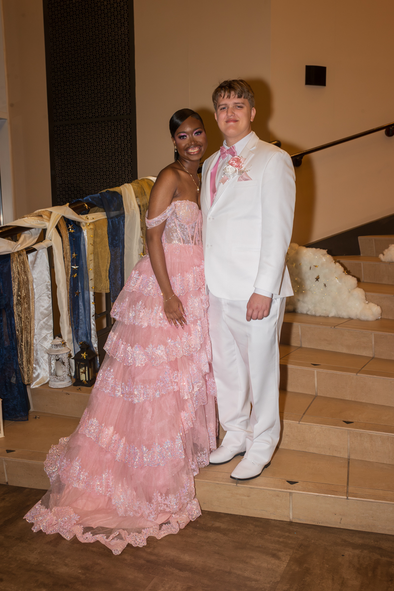 Smiling couple posing on a staircase: woman in pink lace ball gown and a man in a white suit with a pink tie and boutonniere.