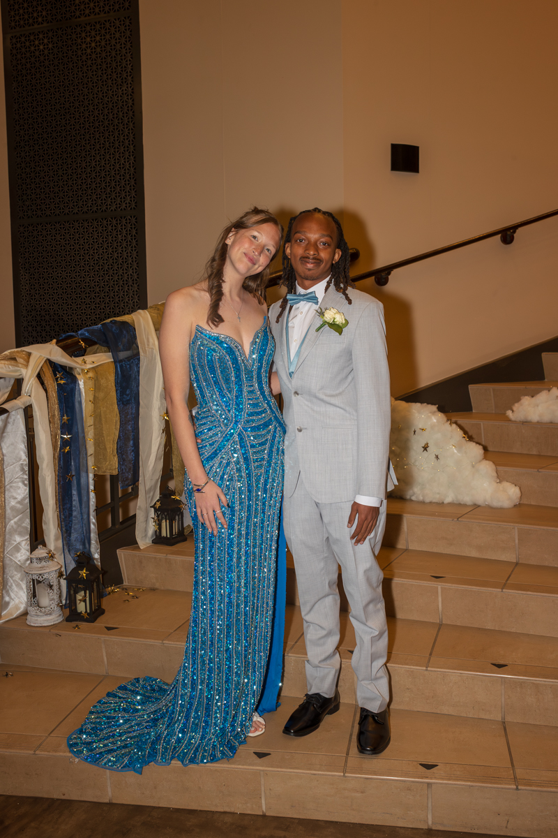 Couple posing on stairs at formal event: woman in a blue sequined gown and man in a light gray suit with bow tie and boutonniere.