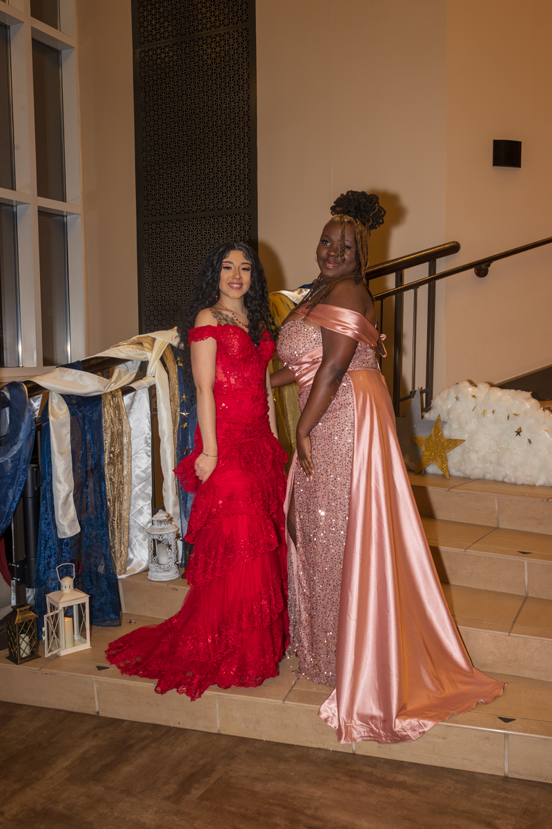 Two women in formal evening gowns pose on a staircase inside a decorated venue: one in a red lace dress and the other in a pink sequined gown with a satin sash, smiling at the camera.