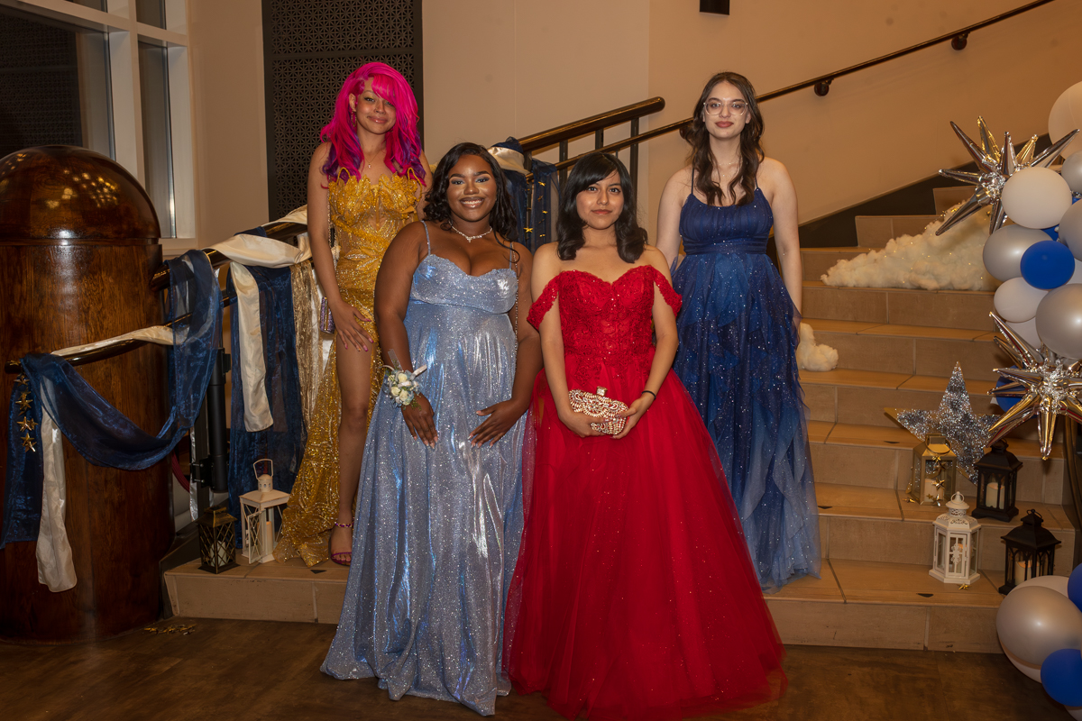 Four young women in glittery gowns pose on a decorated staircase with festive balloons and star accents nearby.