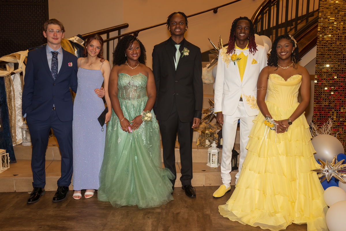 Six teens in formal attire pose on a staircase at a prom-like event: blue suit, lavender dress, green gown, black suit, white suit with yellow accents, and a yellow ballgown.