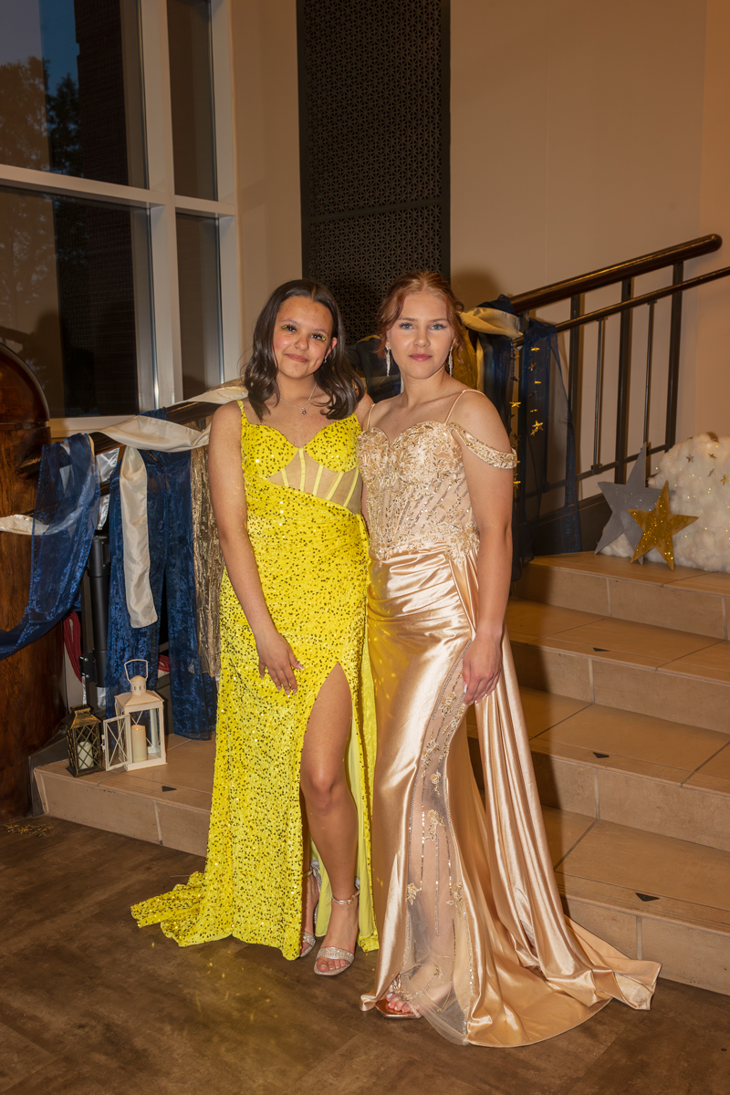 Two young women in glittery evening gowns pose together on a staircase at a decorated indoor event moment.