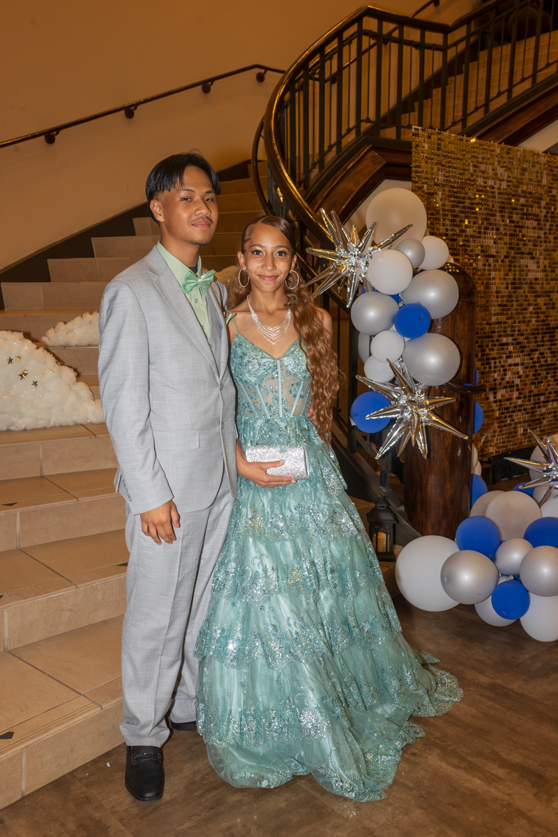 Couple posing on a staircase at a formal event; man in a light gray suit with mint bow tie, woman in a turquoise ball gown holding a silver clutch.