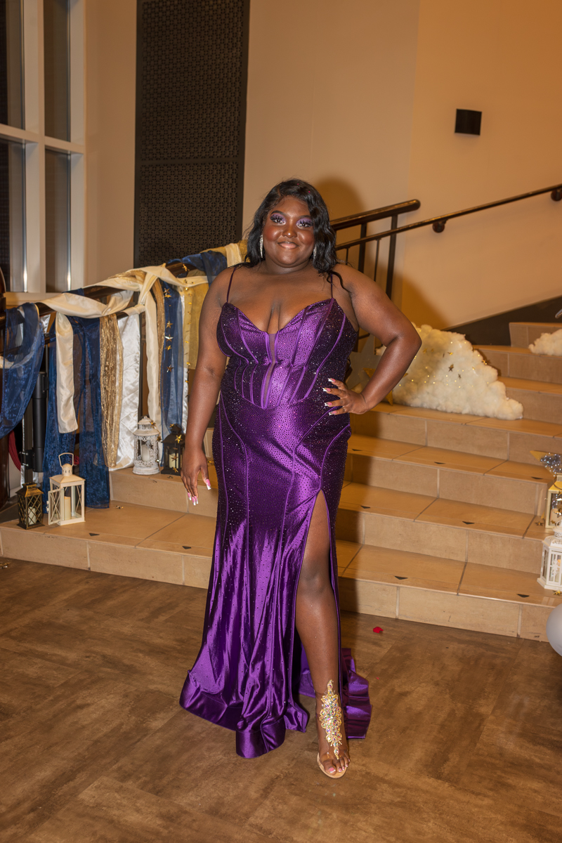 Woman in a shimmering purple gown with a thigh-high slit posing on a staircase at a formal event.