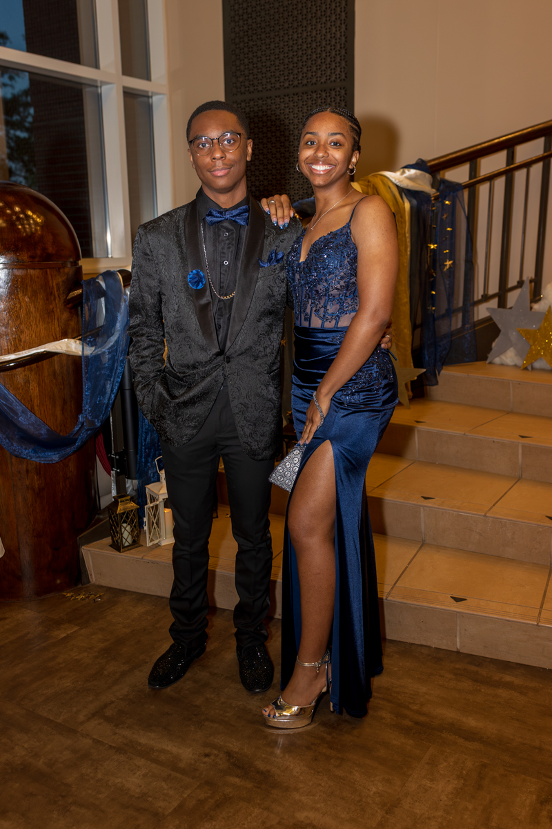 Couple in formal attire posing on a staircase at a party, smiling at the camera. The man wears a black suit and bow tie, the woman a blue embellished gown with a slit.