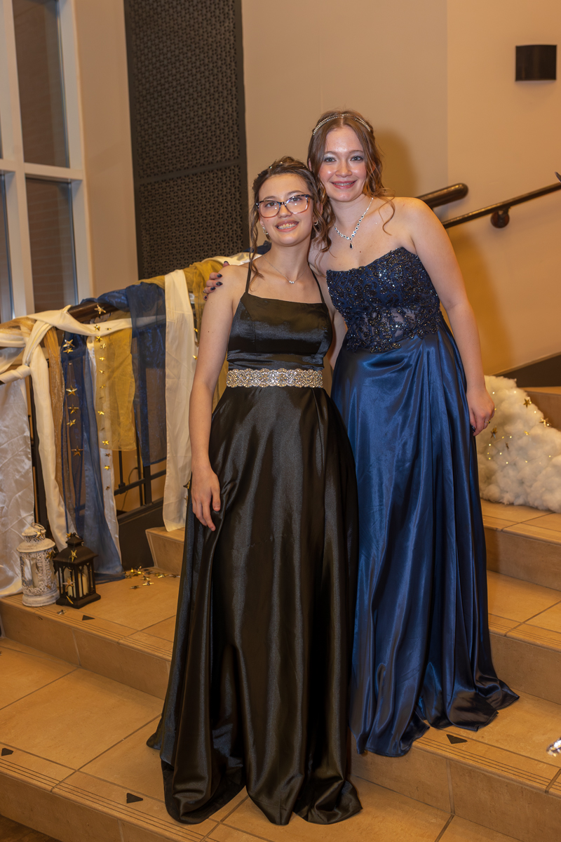 Two young women in formal gowns pose together on a decorated staircase at a formal event; one in a black dress with a silver belt, the other in a blue sequined gown.