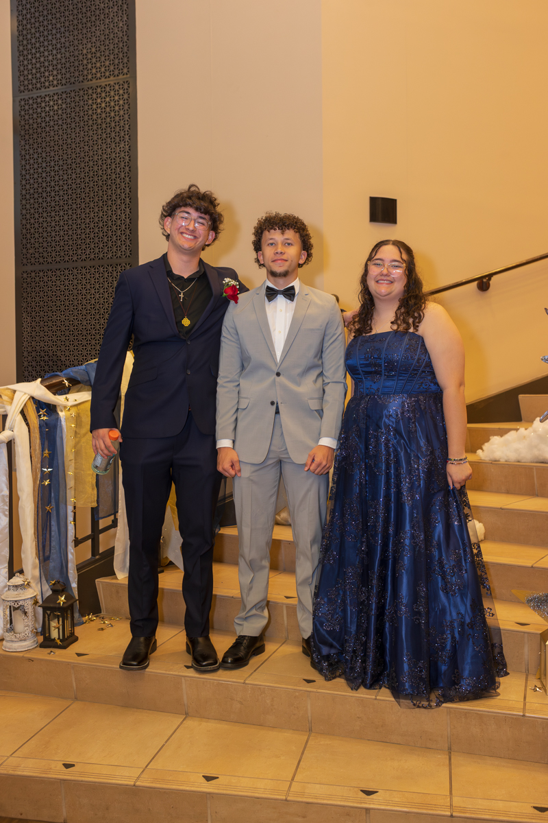 Three friends in formal attire standing on a ramped staircase at a party, smiling for the camera (left: dark suit with boutonniere; center: light gray suit; right: blue gown).
