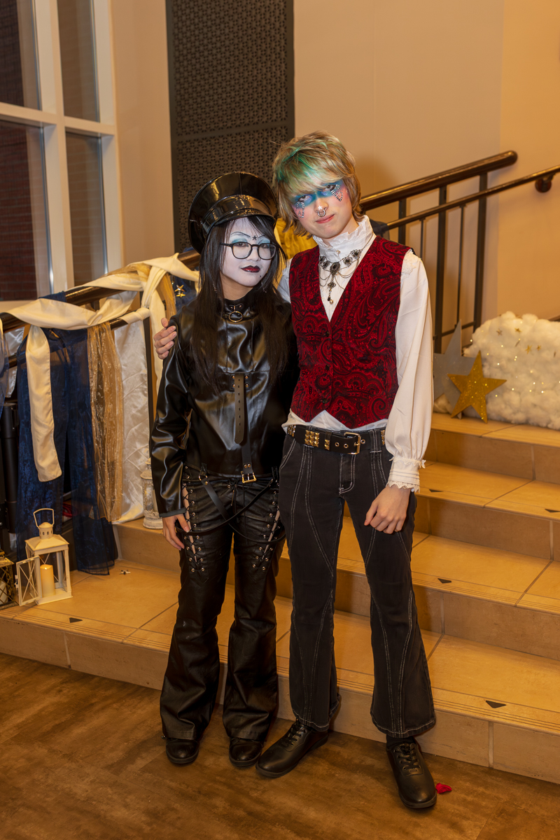 Two costumed individuals pose on a stair landing: one in black leather with a cap and glasses, the other in a white shirt and red velvet vest with blue makeup.