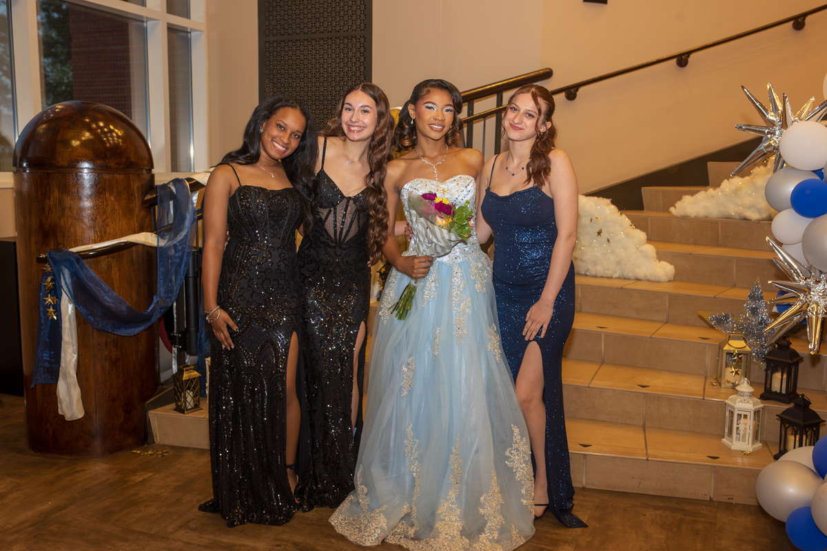 Four young women in formal gowns pose together indoors, the center woman in a light blue ballgown holding a bouquet.