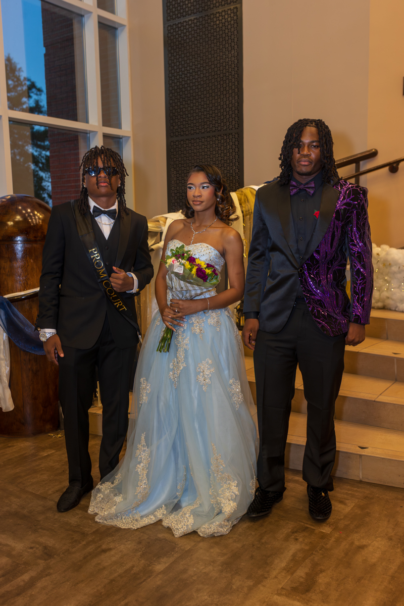 Three students in formal attire pose indoors for prom court photo; the center student wears a light blue gown and holds a bouquet.