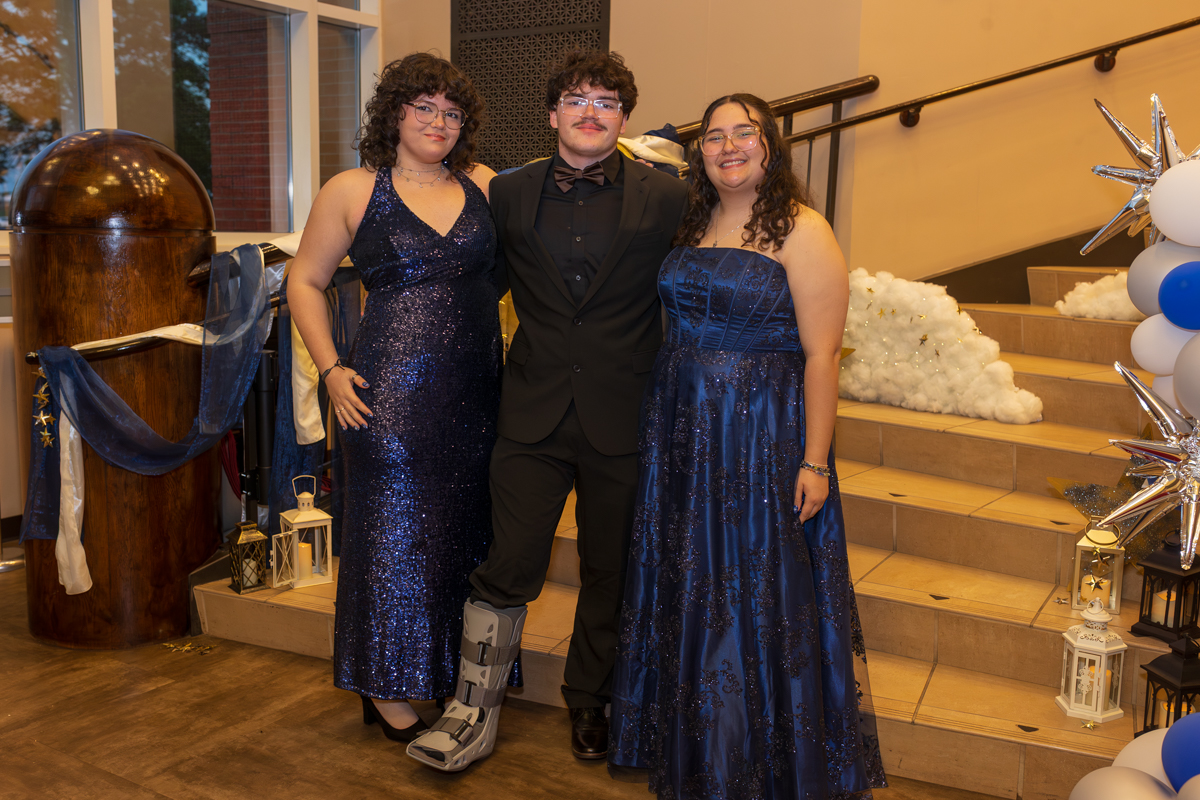 Three friends in formal attire posing together on a decorated staircase at a prom-style event.
