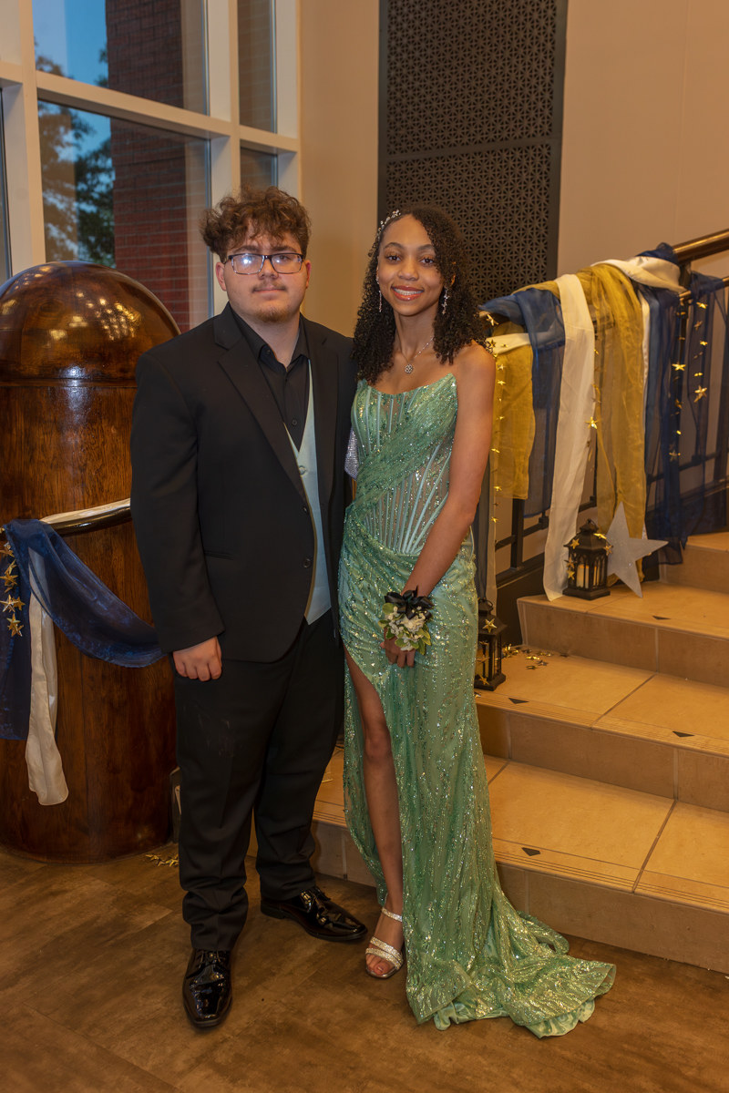 Two young people in formal attire posing together at a decorated indoor event, girl in a teal beaded gown with a slit and corsage smiles beside a young man in a black suit and teal tie.