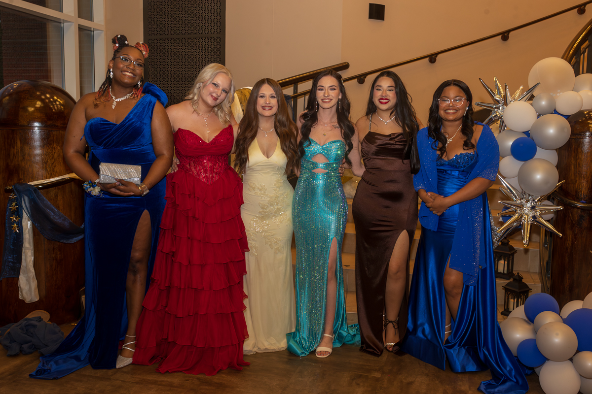Six women in formal gowns pose together on a staircase at a celebration, with balloon decor on the right.