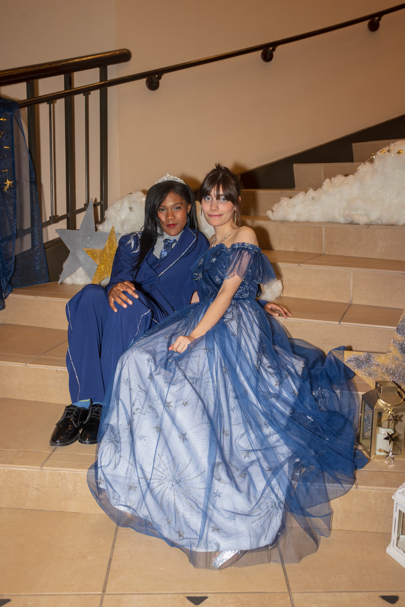 Two teens dressed in blue formal gowns pose together on a stair-step backdrop with star decorations and fake clouds behind them, one wearing a tiara.