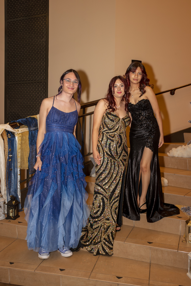 Three young women in formal gowns pose on a stairway at an event: blue layered ball gown, gold-and-black beaded gown, and black sequin dress with slit.