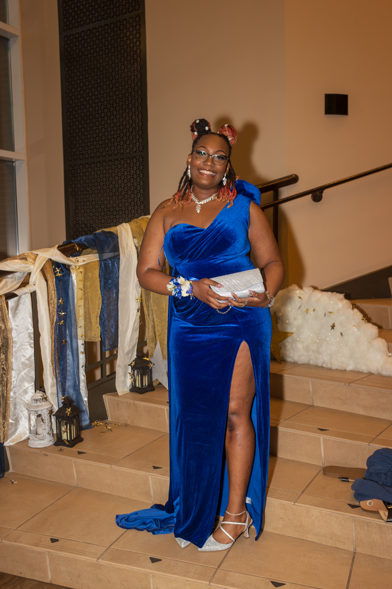 Smiling woman in a royal blue velvet one-shoulder gown with a high slit, posing on a decorated stairway at an event.