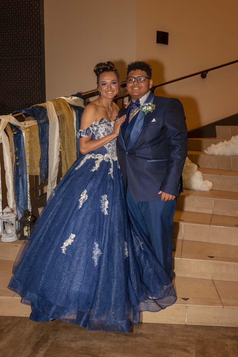 Couple in formal wear posing on a staircase at a special event; the woman wears a navy blue lace ball gown, and the man wears a blue suit with a bow tie and boutonniere.