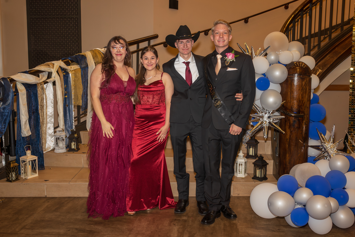 Four friends in formal wear pose together on a staircase, with blue, white, and silver balloon decorations nearby.