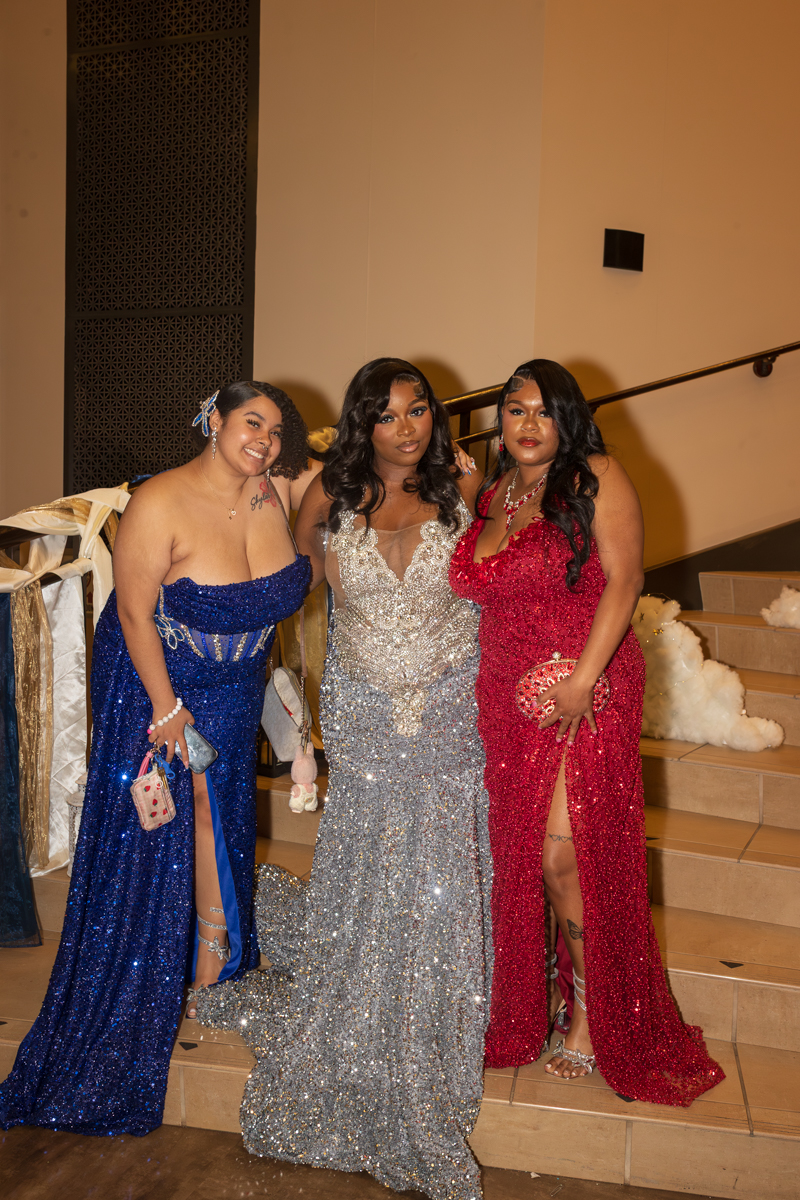 Three women in glittery evening gowns pose on stairs at a formal event; blue, silver, and red dresses sparkle under warm lighting.
