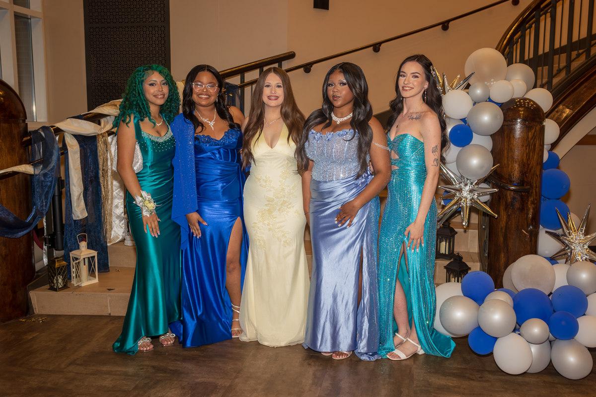 Six young women in formal gowns posing together at an event, with a staircase and balloon decor in the background.