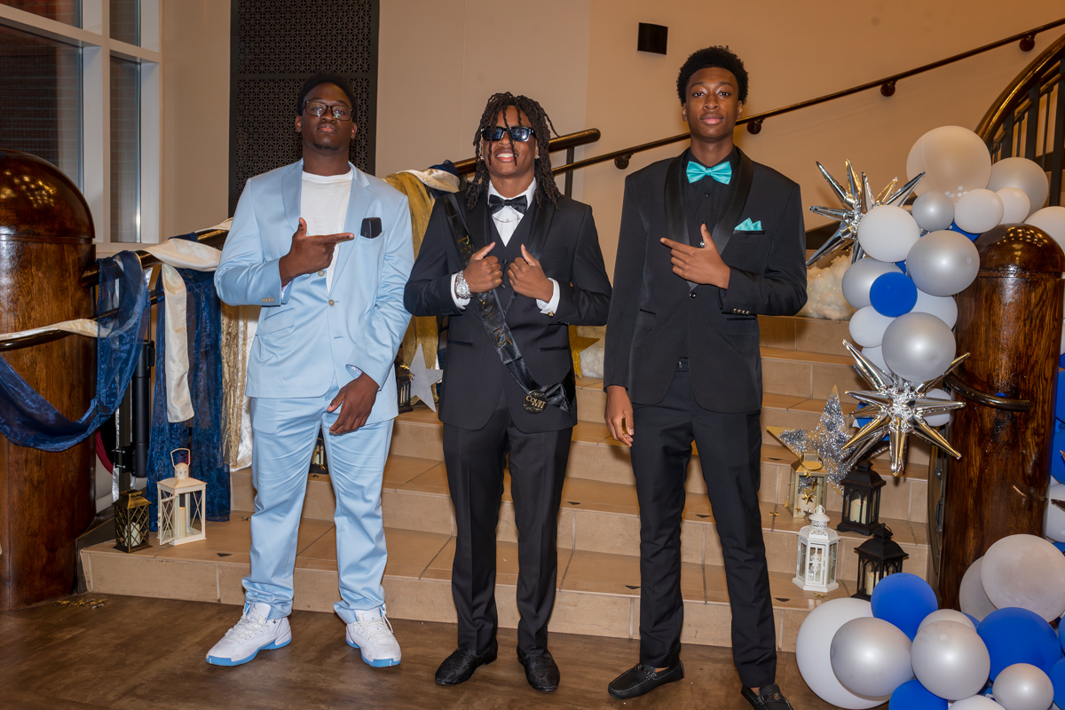 Three young men in formal suits posing on a decorated staircase with balloons and lanterns in blue, white, and silver.