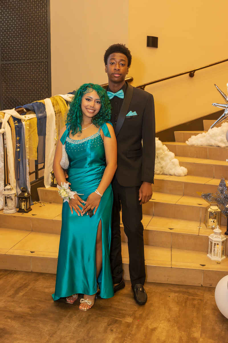 A couple in formal attire posing on a staircase: woman in a teal gown with a thigh-high slit and turquoise accessories, man in a black suit with a turquoise bow tie and pocket square.