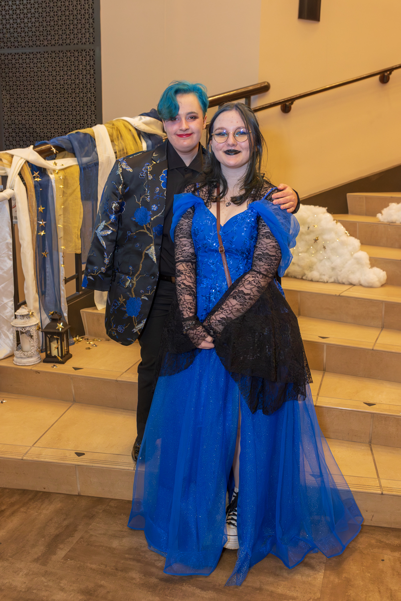 Two friends in costume posing together at a party; one in a blue gown with black lace, the other in a dark floral jacket and teal hair.