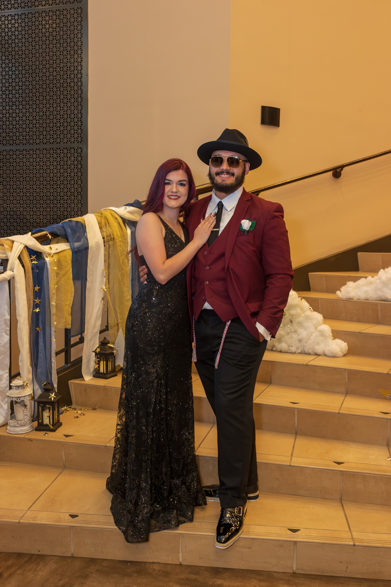 Couple posing on a stairway at a formal event; woman in a black sequin gown and man in a burgundy suit with a hat and sunglasses.