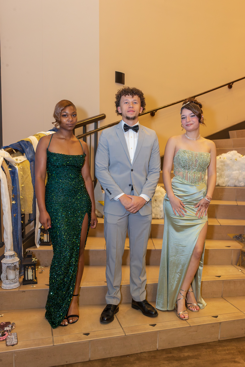 Three teens in formal wear posing on a staircase: a girl in emerald sequin gown, a boy in a light gray suit, and a girl in a pale green dress with a thigh-high slit.