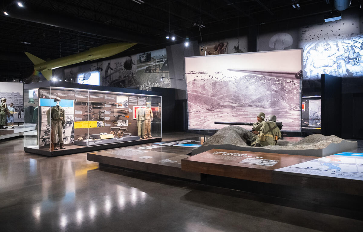 Museum exhibit with glass cases of military uniforms and weapons, and a sand diorama featuring soldiers firing a large gun.