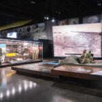 Museum exhibit with glass cases of military uniforms and weapons, and a sand diorama featuring soldiers firing a large gun.