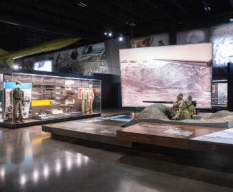 Museum exhibit with glass cases of military uniforms and weapons, and a sand diorama featuring soldiers firing a large gun.