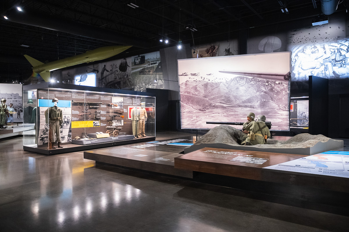 Museum exhibit with glass cases of military uniforms and weapons, and a sand diorama featuring soldiers firing a large gun.