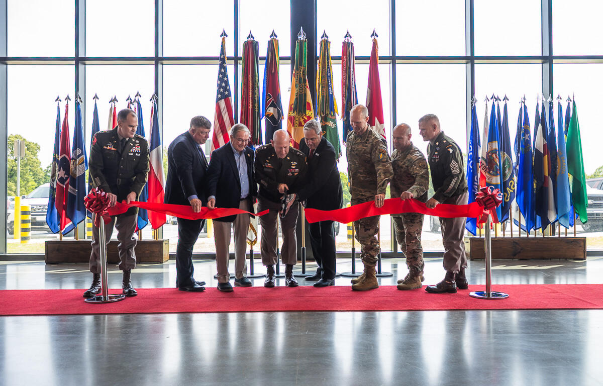 Military personnel and officials cut a red ribbon at a ceremony, with numerous national and military flags behind them inside a glass-walled venue.
