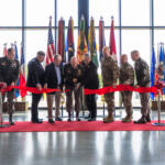 Military personnel and officials cut a red ribbon at a ceremony, with numerous national and military flags behind them inside a glass-walled venue.