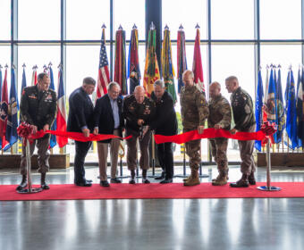 Military personnel and officials cut a red ribbon at a ceremony, with numerous national and military flags behind them inside a glass-walled venue.
