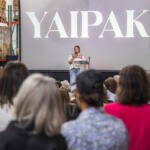 Speaker on stage delivering talk in a warehouse, large 'YAIPAK' logo on screen behind, audience seated in foreground.