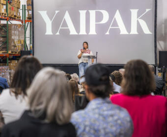 Speaker on stage delivering talk in a warehouse, large 'YAIPAK' logo on screen behind, audience seated in foreground.