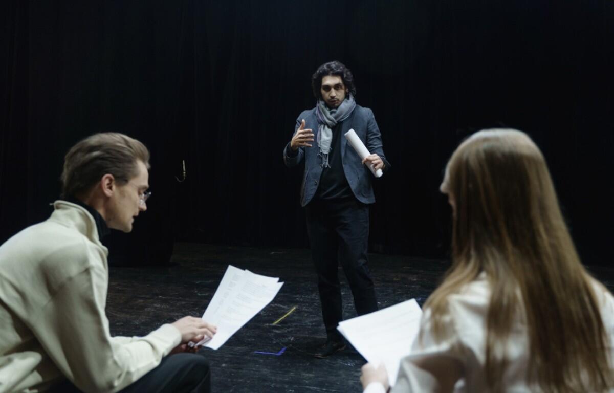 Man in a blazer and scarf speaks to two actors holding scripts in a dim rehearsal space on stage.