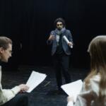 Man in a blazer and scarf speaks to two actors holding scripts in a dim rehearsal space on stage.