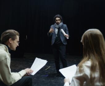 Man in a blazer and scarf speaks to two actors holding scripts in a dim rehearsal space on stage.