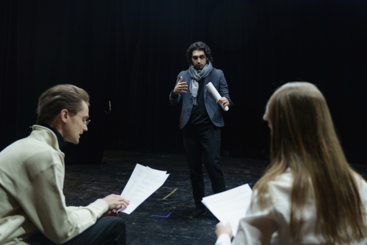 Man in a blazer and scarf speaks to two actors holding scripts in a dim rehearsal space on stage.