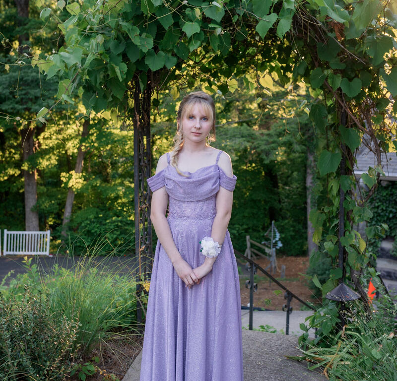 Young woman in a lavender off-shoulder gown stands on a stone path under a leafy arch in a garden, wearing a white corsage on her wrist.